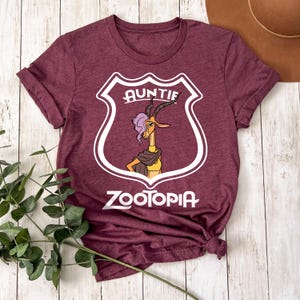 May include: Burgundy t-shirt with a shield design. The word "AUNTIE" is above a cartoon gazelle character with purple hair and a scarf. The word "ZOOTOPIA" is printed below the character in white. The shirt is knotted at the bottom.