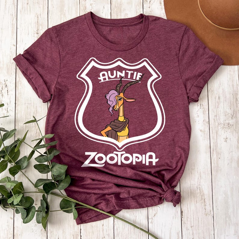 May include: Burgundy t-shirt with a shield design. The word "AUNTIE" is above a cartoon gazelle character with purple hair and a scarf. The word "ZOOTOPIA" is printed below the character in white. The shirt is knotted at the bottom.