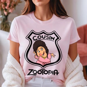 May include: A pale pink t-shirt with a cartoon image of a Zootopia character. The character is framed within a black and white shield, with the word "COUSIN" above and "ZOOTOPIA" below the image.
