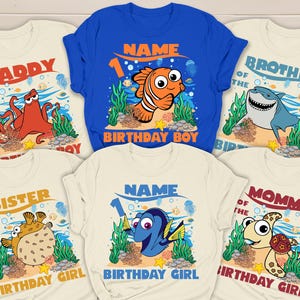 May include: A collection of cream and blue t-shirts with colorful cartoon sea creature graphics. The shirts feature text like "Daddy", "Brother", "Sister", "Mommy", and "Birthday Boy/Girl", with a name and the number "1".