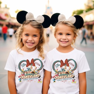 May include: Two young girls wearing black and tan Mickey Mouse ear headbands and white t-shirts. The shirts feature a Disneyland graphic with a bow, castle, and the text "DISNEYLAND FAMILY ADVENTURE EST. 1955".