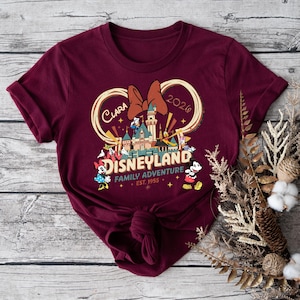 May include: Burgundy t-shirt featuring a Disneyland graphic with a castle, Minnie Mouse bow, and various Disney characters. The text includes "Clara 2026" and "Family Adventure Est. 1955".