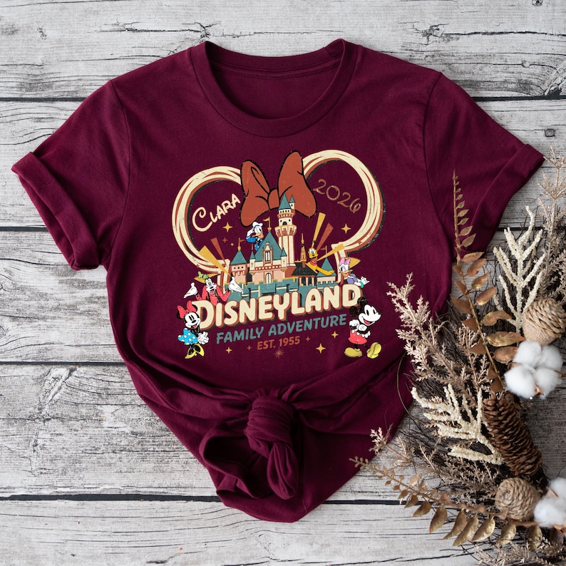 May include: Burgundy t-shirt featuring a Disneyland graphic with a castle, Minnie Mouse bow, and various Disney characters. The text includes "Clara 2026" and "Family Adventure Est. 1955".