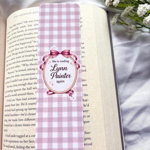 Puede incluir: Un marcapáginas a cuadros rosas y blancos con el texto "She is reading Lynn Painter again" en un óvalo decorativo. El marcapáginas está en un libro abierto. Flores blancas en el fondo.