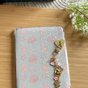 Puede incluir: Una funda transparente para teléfono con un fondo azul claro con un patrón de conchas marinas, estrellas y lazos rosas. Un charm dorado con un corazón, flores, un sombrero de vaquero y un cactus cuelga de la funda.