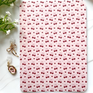 May include: Gold-coloured bow earrings and a pink and gold earring with a pink floral design. The background is a pink surface with a pattern of red cherries and hearts.