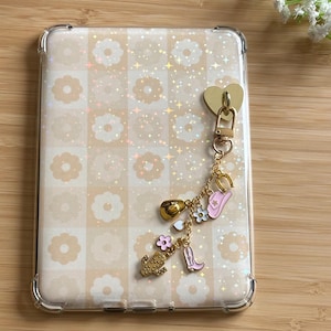 May include: A clear phone case featuring a beige floral checkered design, with a gold heart-shaped charm. The charm has a chain with charms, including a cactus, cowboy boot, and a cowboy hat.