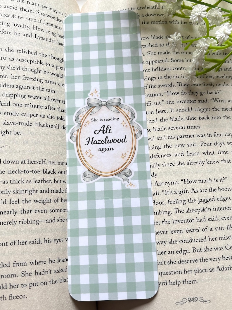 May include: A bookmark with a light green and white gingham pattern. The bookmark features an oval design with the text "She is reading Ali Hazelwood again" in a decorative font, accented with a bow and stars.