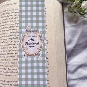 May include: A green and white checkered bookmark with a decorative oval in the center. The oval has the text "She is reading Ali Hazelwood again" in a script font. The bookmark is placed in an open book.