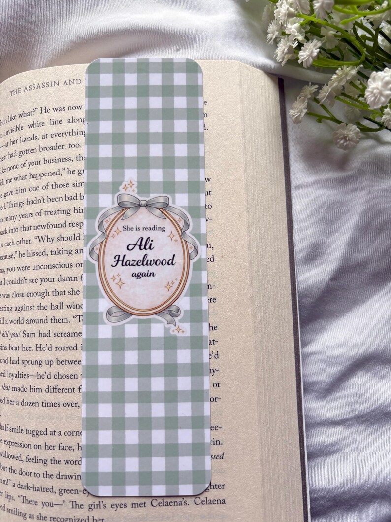 May include: A green and white checkered bookmark with a decorative oval in the center. The oval has the text "She is reading Ali Hazelwood again" in a script font. The bookmark is placed in an open book.