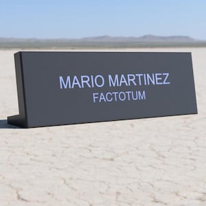 May include: A dark gray desk nameplate with the name "MARIO MARTINEZ" and the title "FACTOTUM" in white lettering. The nameplate is on a cracked, dry surface with a distant mountain range and blue sky.