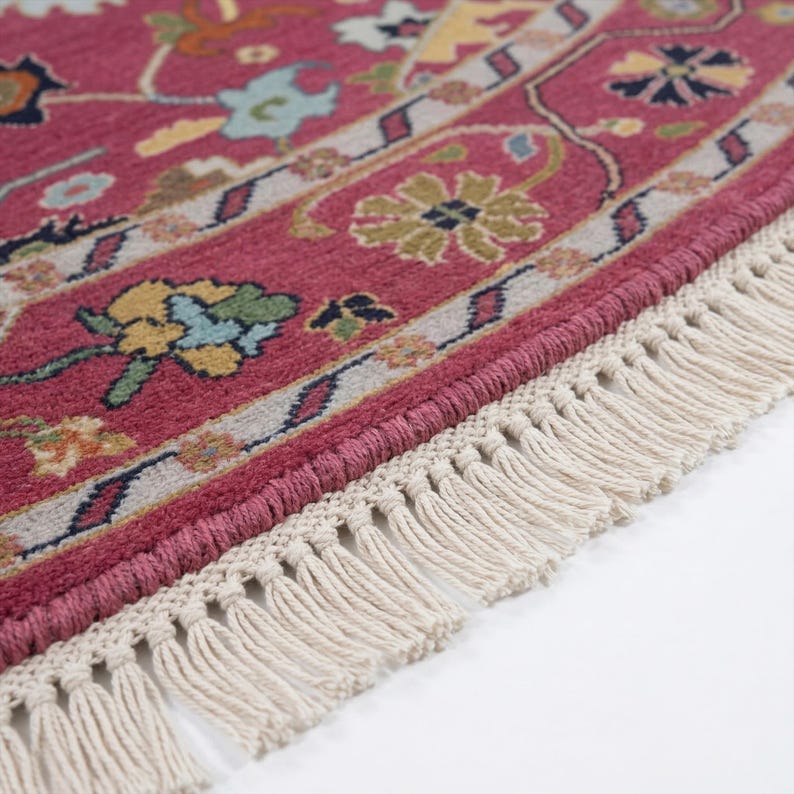 May include: Close-up of a round, red rug with a detailed floral pattern in shades of yellow, blue, and green. The rug has a cream-colored fringe along the edge. The design is intricate and traditional, suitable for home decor.