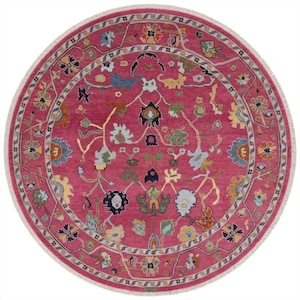 May include: A round rug with a vibrant magenta field and a detailed floral pattern in shades of blue, yellow, green, and white. The rug has a decorative border and a white fringe around the edge. The rug's design is intricate and eye-catching.
