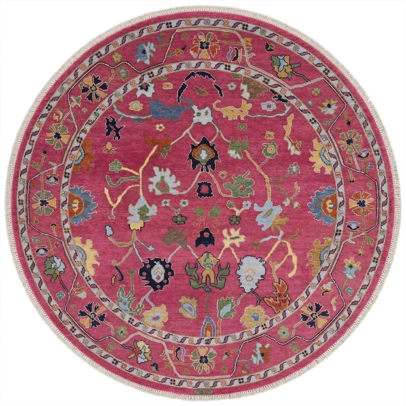 May include: A round rug with a vibrant magenta field and a detailed floral pattern in shades of blue, yellow, green, and white. The rug has a decorative border and a white fringe around the edge. The rug's design is intricate and eye-catching.