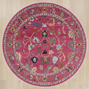 May include: A round, magenta-colored rug with an intricate floral pattern. The rug features a central field of magenta, surrounded by a border with floral and vine motifs in shades of blue, green, yellow, and cream. The rug has a cream-colored fringe.