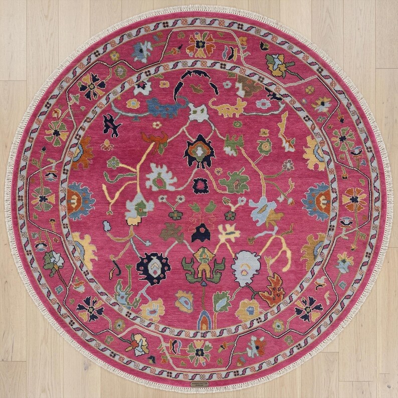 May include: A round, magenta-colored rug with an intricate floral pattern. The rug features a central field of magenta, surrounded by a border with floral and vine motifs in shades of blue, green, yellow, and cream. The rug has a cream-colored fringe.