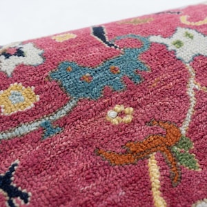 May include: Close-up of a section of a rug with a deep pink background. The rug features a repeating pattern of teal, yellow, and orange floral and geometric designs. The texture of the rug is visible, with a slightly raised pile.