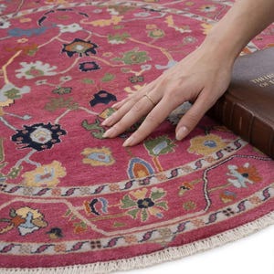 May include: A round, pink rug with a detailed floral pattern in shades of blue, green, yellow, and white. The rug has a fringed edge and is partially covered by a hand and a brown book.