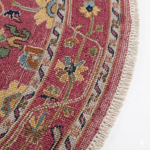 May include: Close-up of a round, antique rug with a detailed floral pattern. The rug features a deep red background with yellow, blue, and green floral motifs. The border has a cream-colored fringe.