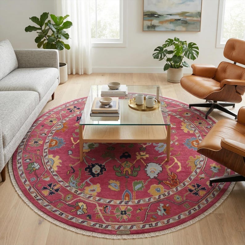 May include: A round, magenta-colored rug with an intricate floral pattern and a cream-colored fringe. The rug is centered in a living room setting, with a glass-top coffee table and leather chairs.