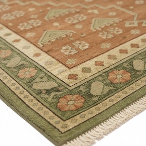 May include: Detailed shot of a patterned rug in warm tones. The central section is terracotta, with geometric and floral designs in beige and green. The border features floral patterns, and the rug has a fringed edge.