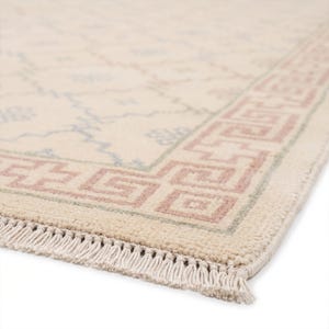 Könnte beinhalten: Ein cremefarbener Teppich mit einem hellblauen geometrischen Rautenmuster in der Mitte und einer rosa Bordüre im griechischen Schlüsseldesign. Der Teppich hat eine kurzflorige Textur und ist mit einer Fransenborte aus cremefarbenen Quasten versehen.