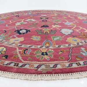 May include: A round, pink rug with an intricate floral pattern. The rug features a detailed border and a fringe along the edge. The design includes various colors like blue, yellow, and green, creating a vibrant and decorative piece.