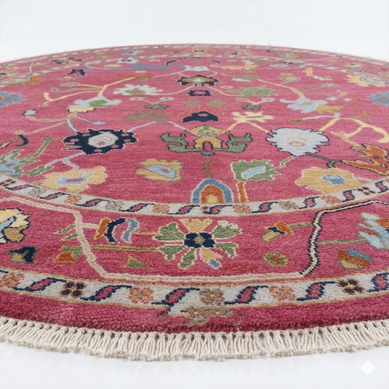 May include: A round, pink rug with an intricate floral pattern. The rug features a detailed border and a fringe along the edge. The design includes various colors like blue, yellow, and green, creating a vibrant and decorative piece.