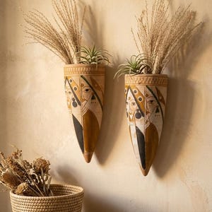 May include: Two wall-mounted planters with a painted face design, filled with dried grasses and small green plants. The planters are beige, brown, and black, and are mounted on a light beige wall. A woven basket and a small dish with stones are on a wooden surface.