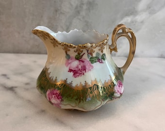 May include: A porcelain creamer featuring a white base with pink rose floral patterns and a deep green lower section. The piece is accented with intricate gold filigree detailing along the scalloped rim and handle, showcasing a curved spout.