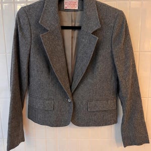 Puede incluir: Una blazer gris con cierre de un solo botón, dos bolsillos delanteros y solapa con muescas. La blazer está colgada de una percha negra. La etiqueta dice "Pendleton".