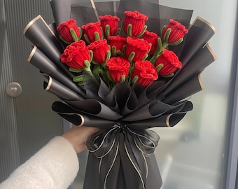 Könnte beinhalten: Ein Strauß aus gehäkelten roten Rosen mit grünen Stielen und Blättern, eingewickelt in Lagen aus schwarzem Papier mit goldfarbenen Rändern. Das Arrangement ist an der Basis mit einem schwarzen, transparenten Band mit feiner Goldkante gebunden.