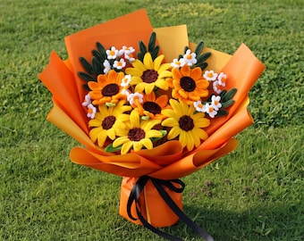 Könnte beinhalten: Ein Blumenstrauß aus gehäkelten Sonnenblumen und kleinen weißen Blüten mit dunkelgrünen Blättern, eingewickelt in leuchtend orangefarbenes Papier und mit einem schwarzen Band gebunden. Die Anordnung zeigt gelbe Blütenblätter und dunkelbraune Mitten.
