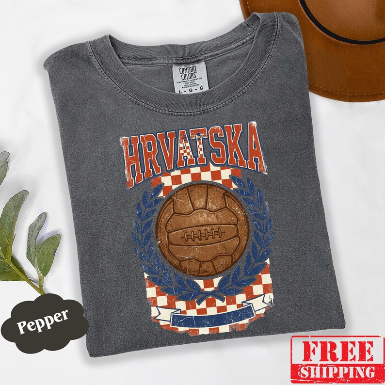 May include: A charcoal gray t-shirt featuring a vintage-style graphic with the text HRVATSKA in red and white. The design includes a brown soccer ball, a red and white checkered pattern, and blue laurel leaves. The shirt has a crew neck collar.