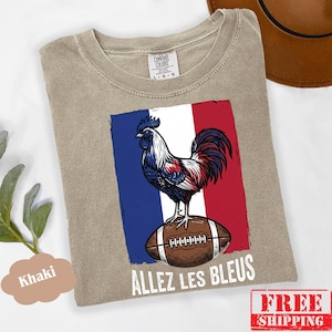 Puede incluir: Una camiseta de cuello redondo color caqui con un gr&aacute;fico de un gallo de pie sobre un bal&oacute;n de f&uacute;tbol americano marr&oacute;n sobre un fondo de bandera azul, blanca y roja. El texto "ALLEZ LES BLEUS" est&aacute; impreso en blanco bajo el gr&aacute;fico.