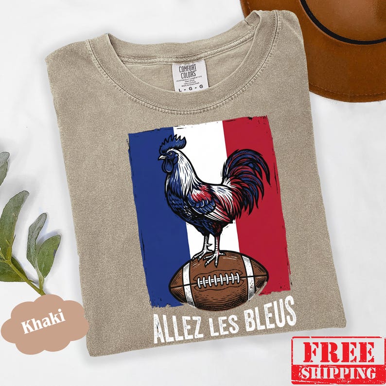 Puede incluir: Una camiseta de cuello redondo color caqui con un gr&aacute;fico de un gallo de pie sobre un bal&oacute;n de f&uacute;tbol americano marr&oacute;n sobre un fondo de bandera azul, blanca y roja. El texto "ALLEZ LES BLEUS" est&aacute; impreso en blanco bajo el gr&aacute;fico.