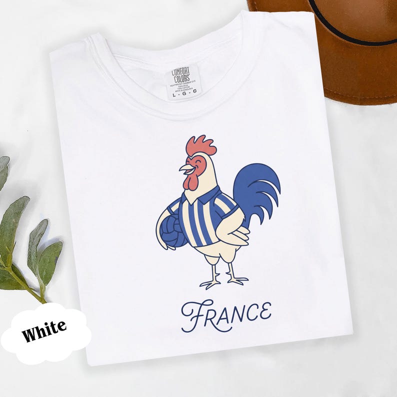 May include: White t-shirt with a cartoon rooster wearing a blue and white striped jersey and holding a football. The word "France" is written in a cursive font below the rooster.