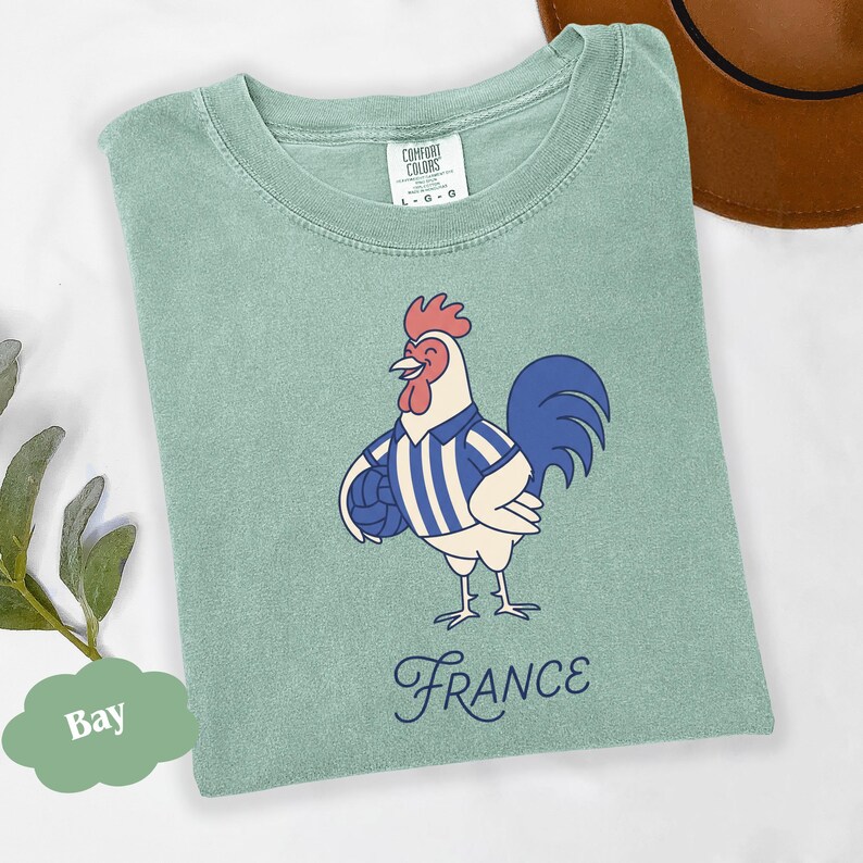 May include: A pale green Comfort Colors t-shirt with a cartoon rooster wearing a blue and white striped shirt and holding a football. The word "France" is written in a cursive font below the rooster.