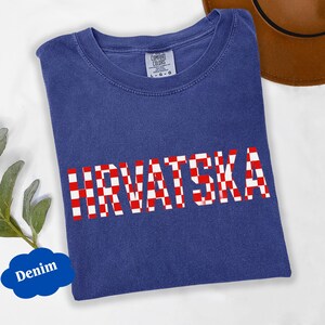 Może przedstawiać: Niebieska koszulka dżinsowa z napisem "HRVATSKA" w czerwono-białą kratę. Koszulka ma okrągły dekolt i kr&oacute;tkie rękawy. Widoczna jest metka marki Comfort Colors.