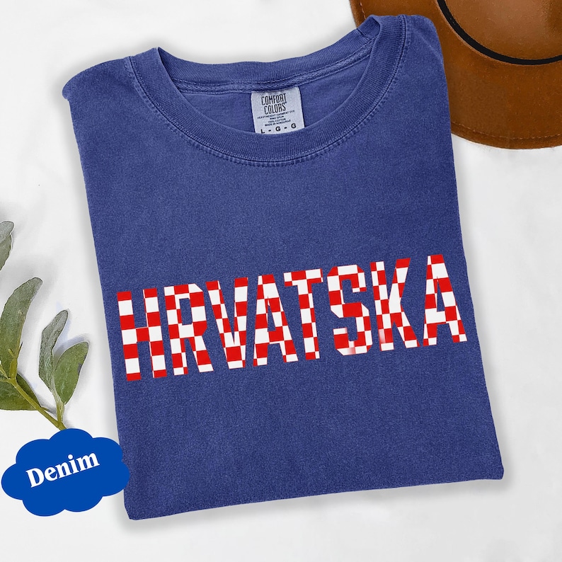 Może przedstawiać: Niebieska koszulka dżinsowa z napisem "HRVATSKA" w czerwono-białą kratę. Koszulka ma okrągły dekolt i kr&oacute;tkie rękawy. Widoczna jest metka marki Comfort Colors.