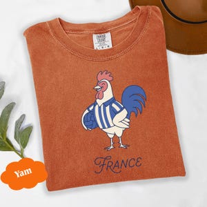 May include: Burnt orange t-shirt with a cartoon rooster wearing a blue and white striped shirt and holding a football. The word "France" is written in a cursive font below the rooster. The shirt is a Comfort Colors brand.