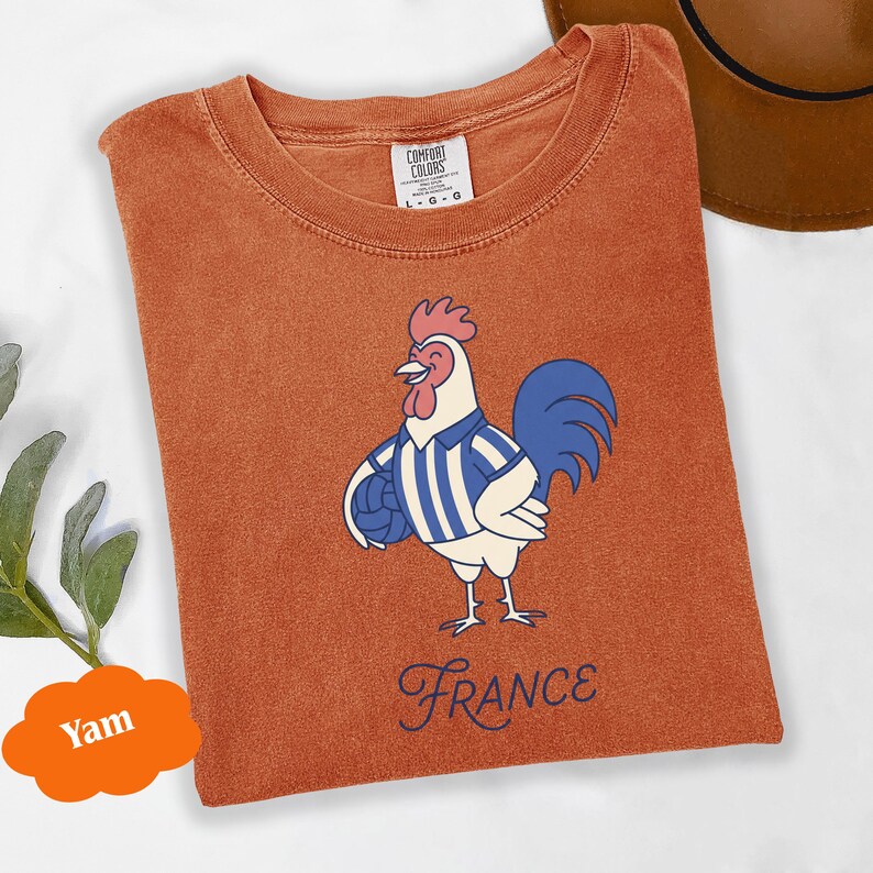 May include: Burnt orange t-shirt with a cartoon rooster wearing a blue and white striped shirt and holding a football. The word "France" is written in a cursive font below the rooster. The shirt is a Comfort Colors brand.