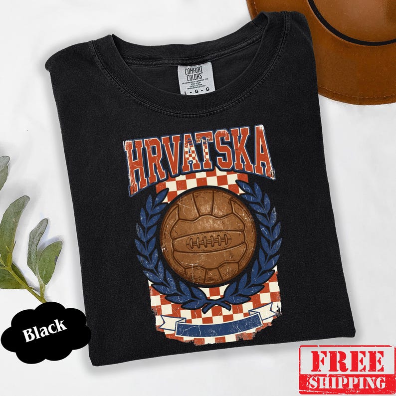 May include: A black short-sleeve t-shirt featuring a graphic print with the text HRVATSKA in red and white. The design includes a brown vintage-style soccer ball centered over a red and white checkered pattern, framed by blue laurel leaves.