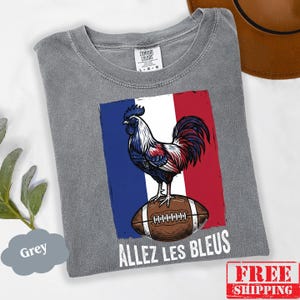 Puede incluir: Una camiseta gris de manga corta con un gr&aacute;fico de un gallo de pie sobre un bal&oacute;n de f&uacute;tbol americano marr&oacute;n sobre un fondo de bandera azul, blanca y roja. El texto "ALLEZ LES BLEUS" est&aacute; impreso en blanco debajo del gr&aacute;fico. Etiqueta Comfort Colors.