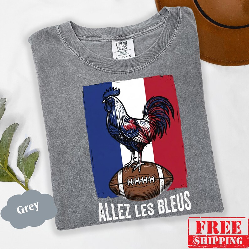 Puede incluir: Una camiseta gris de manga corta con un gr&aacute;fico de un gallo de pie sobre un bal&oacute;n de f&uacute;tbol americano marr&oacute;n sobre un fondo de bandera azul, blanca y roja. El texto "ALLEZ LES BLEUS" est&aacute; impreso en blanco debajo del gr&aacute;fico. Etiqueta Comfort Colors.