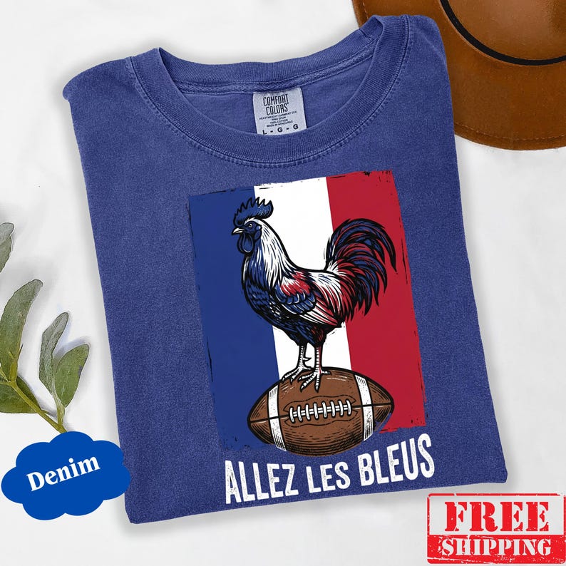 Puede incluir: Una camiseta azul denim con un gr&aacute;fico de un gallo de pie sobre un bal&oacute;n de f&uacute;tbol americano marr&oacute;n sobre un fondo con los colores de la bandera francesa. El texto "ALLEZ LES BLEUS" est&aacute; impreso en blanco debajo del bal&oacute;n. Etiqueta Comfort Colors.