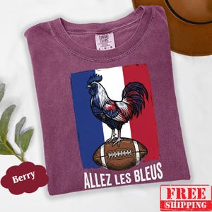 Puede incluir: Una camiseta color baya con un gr&aacute;fico de un gallo de pie sobre un bal&oacute;n de f&uacute;tbol americano marr&oacute;n frente a la bandera francesa. El texto "ALLEZ LES BLEUS" est&aacute; impreso en blanco debajo del gr&aacute;fico. La camiseta tiene una etiqueta Comfort Colors.
