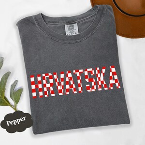 Może przedstawiać: Ciemnoszara koszulka z napisem "HRVATSKA" w czerwono-białą kratę. Koszulka ma okrągły dekolt i kr&oacute;tkie rękawy. Brązowy kapelusz jest widoczny w prawym g&oacute;rnym rogu.