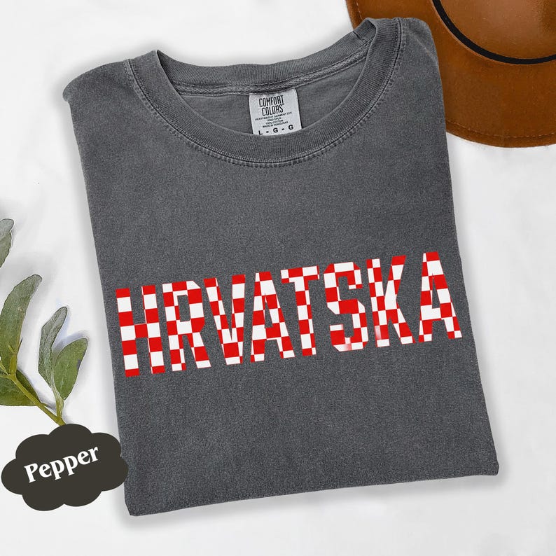 Może przedstawiać: Ciemnoszara koszulka z napisem "HRVATSKA" w czerwono-białą kratę. Koszulka ma okrągły dekolt i kr&oacute;tkie rękawy. Brązowy kapelusz jest widoczny w prawym g&oacute;rnym rogu.