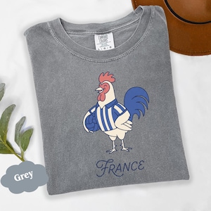 May include: A grey t-shirt with a cartoon rooster wearing a blue and white striped shirt and holding a ball. The word "France" is written in cursive below the rooster. The shirt is labeled "Comfort Colors".